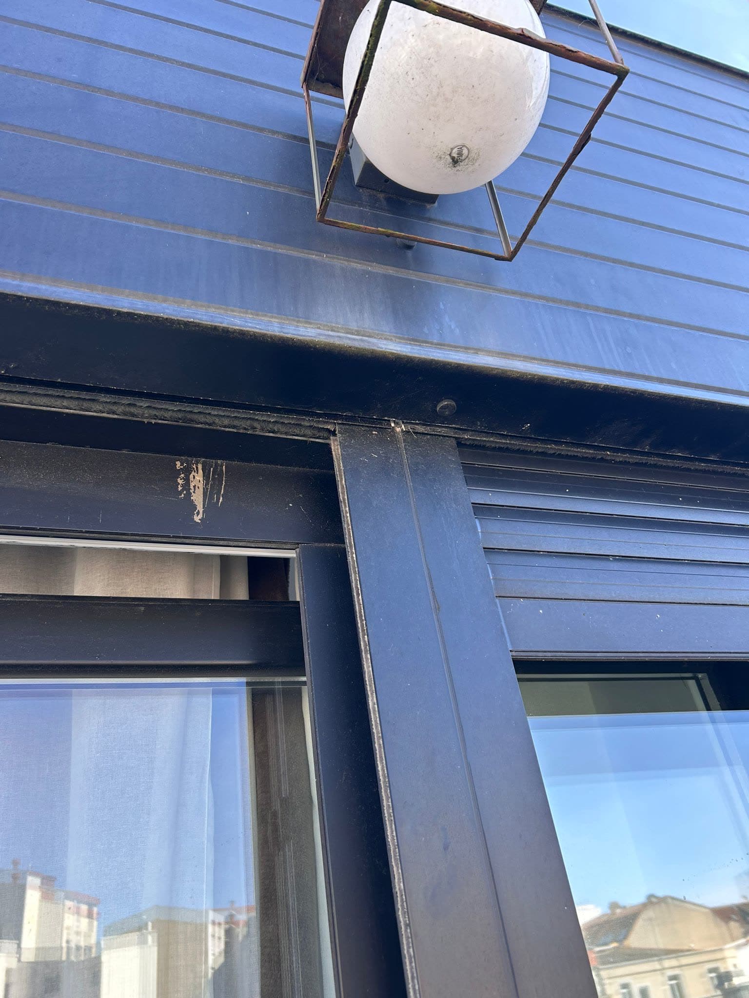 Vista desde la calle del cajón exterior de una persiana sobre una ventana de carpintería oscura, con el paño recogido dentro del cajón y un aplique de pared al lado
