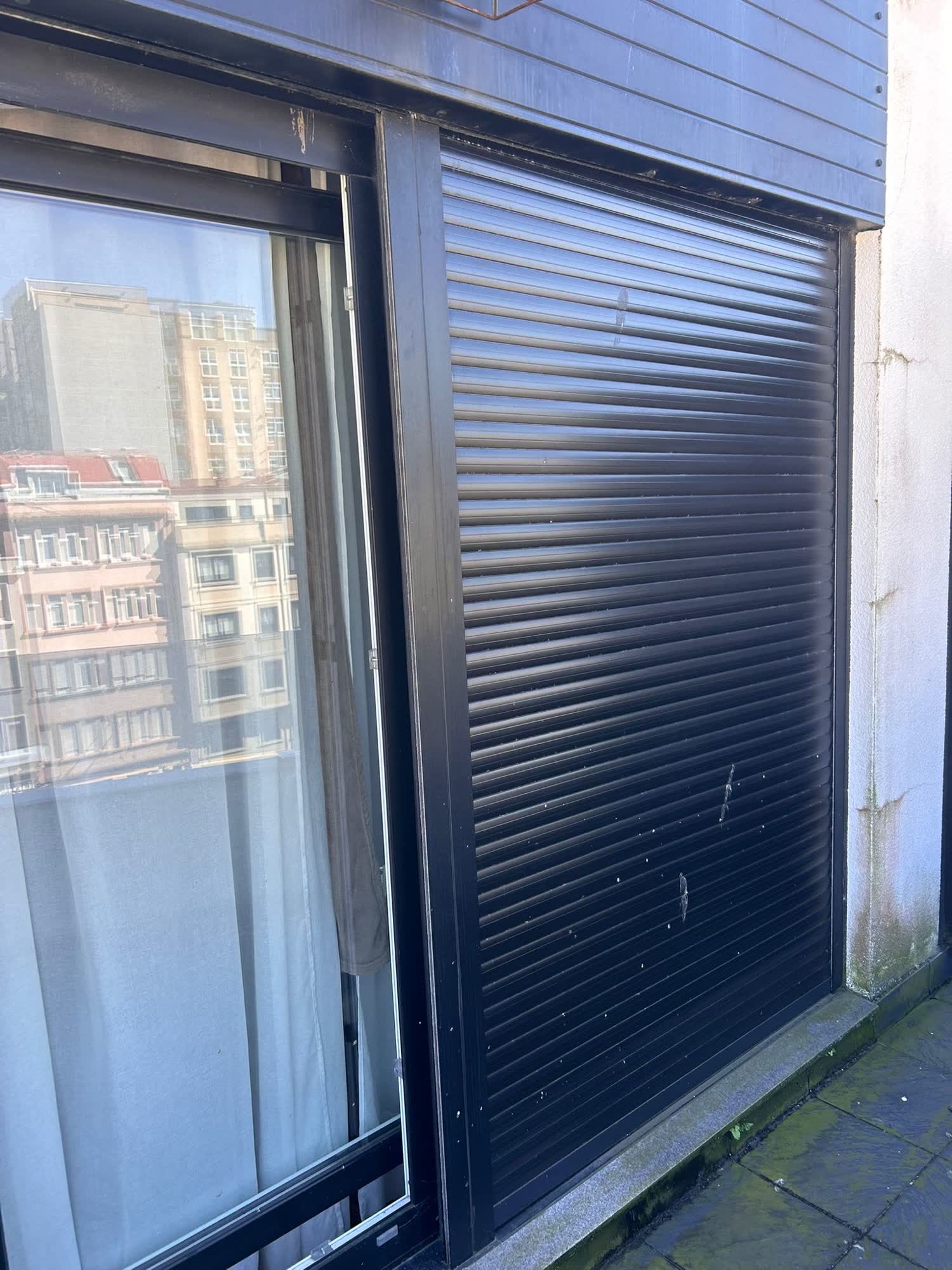 Persiana oscura bajada hasta el suelo en el hueco de una terraza, junto a la hoja corredera abierta que refleja los edificios de enfrente