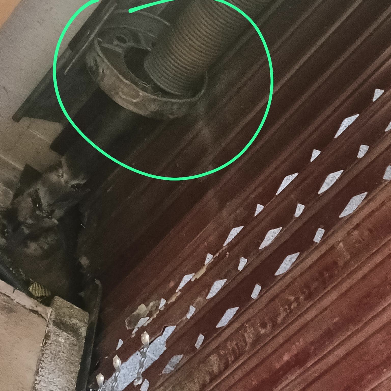 Primer plano del eje de una persiana enrollable con el muelle de recuperación rodeado por un círculo verde dibujado sobre la foto, con las lamas marrones manchadas de pintura debajo