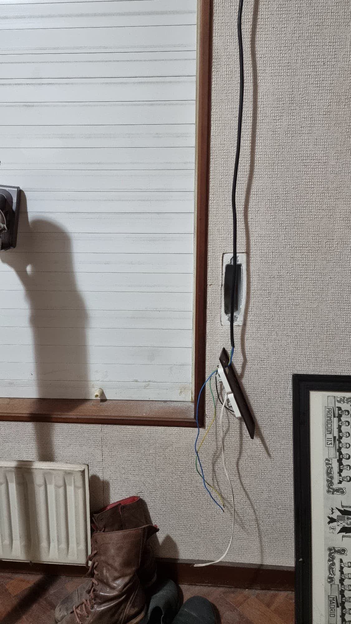 Instalación del mando de una persiana motorizada en una vivienda: hueco del antiguo recogedor abierto en la pared, cable de alimentación bajando y el pulsador descolgado con los conductores sin conectar