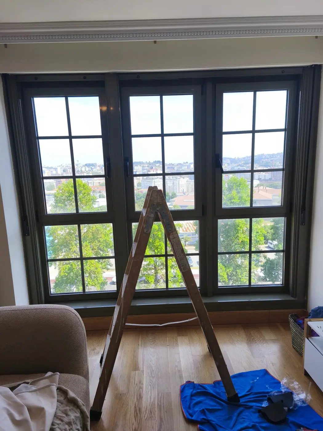 Salón de una vivienda con un ventanal de tres hojas de carpintería verde oscuro y una escalera de tijera abierta delante, con la lona de protección extendida sobre el parqué durante el montaje