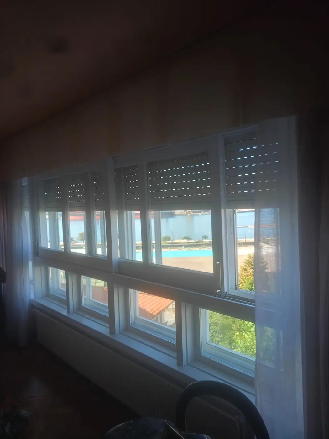 Galería acristalada a contraluz con las persianas de lama microperforada bajadas a media altura sobre las ventanas correderas blancas